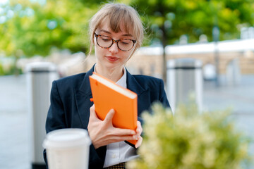 Young woman in glasses reading an orange notebook while enjoying coffee at an outdoor cafe on a sunny day in the city