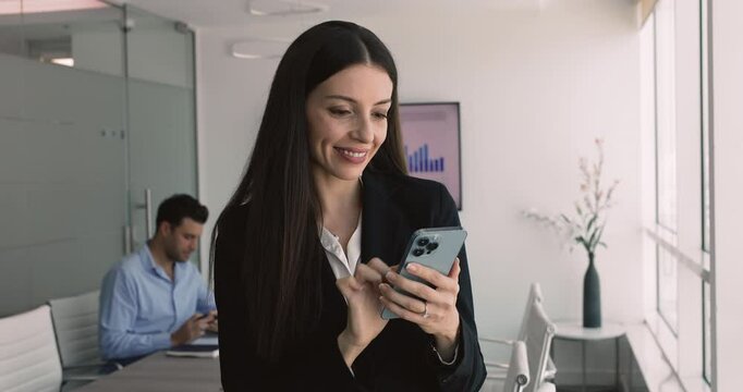 Businesswoman using her smartphone while standing in modern workplace, checking messages, emails, or business-related information, responding to important notification or managing work tasks remotely