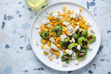 Plate with corn, grilled Brussels sprouts and cheese, horizontal shot on a white and blue granite background, high angle view