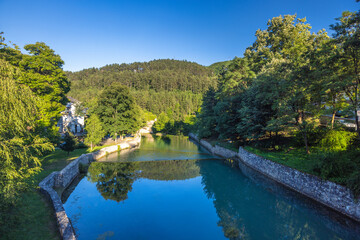 Fototapeta premium The Pliva river in Jajce town in Bosnia and Herzegovina, Europe. Scenic river view with stone embankments surrounded by lush green trees and a forested hillside under a clear blue sky.