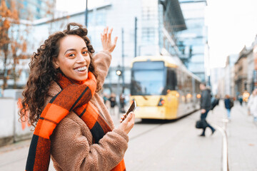 Young woman waves goodbye at a tram stop in modern city during sunny afternoon, demonstrating a joyful farewell
