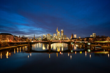 Obraz premium Frankfurt cityscape at night, with illuminated skyscrapers reflecting on the Main River, a lit bridge, and a clear dark blue sky, showcasing urban beauty and tranquility.