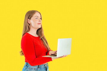 Young woman works on laptop against bright yellow background while standing in casual attire and focusing on her task