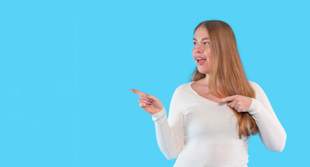 Woman with long hair smiling and pointing towards a blue background while dressed in a casual white...