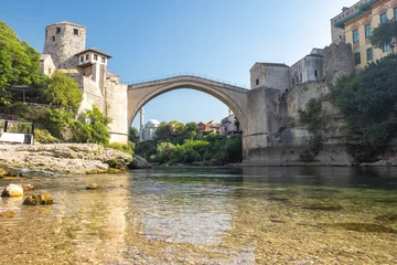 The Stari Most bridge, old bridge on Neretva river in Mostar town, Bosnia and Herzegovina, Europe. Arching stone bridge spans a tranquil river, connecting ancient structures under a clear sky.