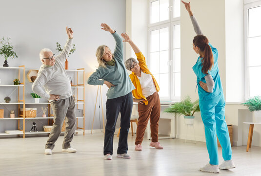 Female nurse or physiotherapist doing healthy stretching exercises for wellness and health with a group of seniors. Elderly people working out in fitness class of nursing home with instructor.