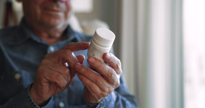 Close up cropped view older retired man sitting in armchair holding and carefully examining small bottle of medicine or supplements, reading label for dosage instructions. Health awareness, self-care