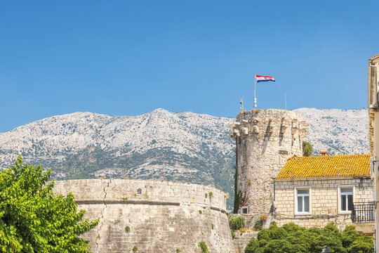 Korcula town in island of Korcula in Adriatic sea in Croatia, Europe. Picturesque coastal town featuring a stone fortress tower topped with a flag, backed by mountainous landscape under a blue sky.