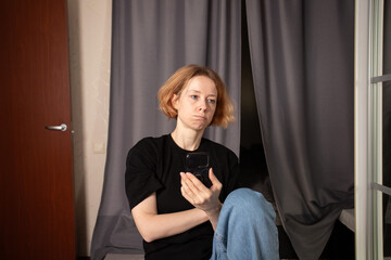 Sad woman with smartphone at home, expressing sorrow and concern about online bullying, dealing...