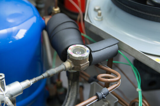 Close-up of sight glass in refrigeration system with insulated copper pipes and sensor wiring, used for monitoring refrigerant flow and diagnostics in HVAC or cooling equipment.