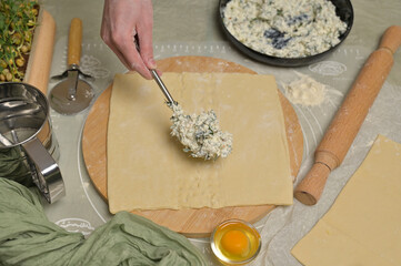Woman's hand with fork putting cottage cheese with parsley and dill greens on dough. Rolling pin, cutter, egg, green towel and microgreens sprouts on kitchen table. Home cooking baking puffs