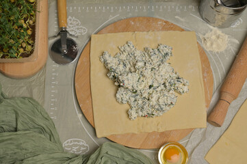 Cottage cheese with parsley and dill greens on dough. Rolling pin, cutter, egg, green towel and microgreens sprouts on kitchen table. Home cooking baking puffs