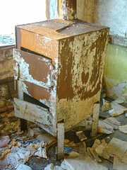 Image depicts a dilapidated interior space, centered around an old, rusted metal storage unit with peeling paint The floor is strewn with debris, and the background shows more decaying structures N