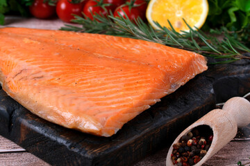 Hot smoked trout fillet on a wooden board. Close-up. Lettuce, cherry tomatoes, lemon, rosemary and pepper are arranged around