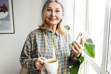 Senior lady in casual shirt with cup of coffee enjoying morning holding smartphone in hands, browsing news feed, standing in front of window and looking at camera with happy facial expression