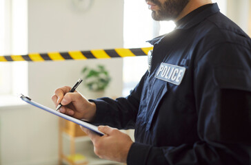 Close up portrait of confident male police officer in uniform standing at fenced crime scene making notes during investigation. Young policeman working indoors. Robbery concept.