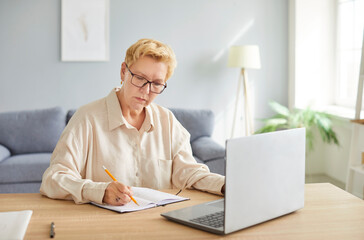 Senior busy mature woman wearing glasses writing notes, using laptop computer, sitting at table at...