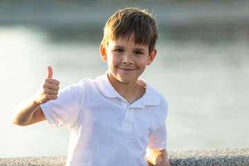 A little boy in casual summer clothes stands on the city embankment along the river, gazes into the camera and shows thumbs up at sunset. Copy space. Childhood, happiness concept.