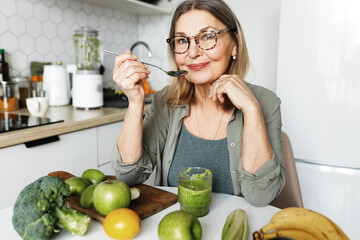 Healthy eating. Secrets of longevity. Senior lady in glasses enjoying breakfast at home, sitting at kitchen table with spoon in hand, having home-made green smoothie prepared form organic fruits