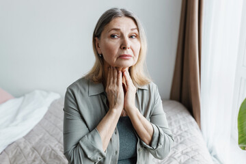 Indoor portrait of worried senior woman siting on bed in bedroom examining her thyroid, touching...