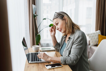 Side view of puzzled senior businesswoman working remotely from home, checking mailbox, looking at screen touching her forehead thinking over appropriate reply on e-mail from partners
