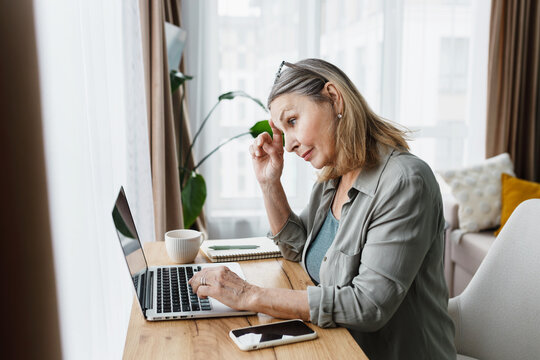 Side view of puzzled senior businesswoman working remotely from home, checking mailbox, looking at screen touching her forehead thinking over appropriate reply on e-mail from partners