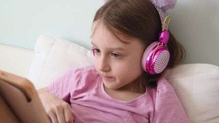 Worried girl wearing bright pink headphones bites finger anxiously while looking at tablet screen, sitting nervously on bed at home.