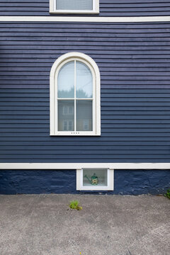 The exterior wooden wall of a vintage blue house with white painted trim. The building has three differently sized windows of various styles. The middle window is a half-round with four glass panes.
