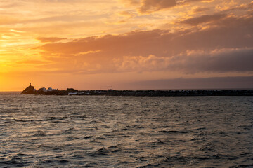 A dramatic orange sky at sunset with fluffy clouds. There's a breakwater wall with a nautical navigation light in the distance. The horizon is golden over the ocean. The rock wall protects the harbor.