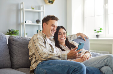 Young married couple with mobile phone sitting on sofa together. Happy man and woman relaxing on...