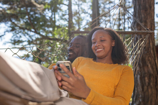 African American couple relaxing on a hammock, scrolling on smartphones, and enjoying nature outdoors in a glamping site in Europe.