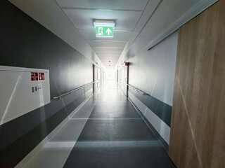 Long and empty hospital corridor with bright lights from window and green evacuation exit sign in the foreground