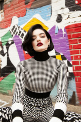 Stylish woman with retro hair and vibrant makeup poses in black and white pattern clothing against a colorful graffiti background.