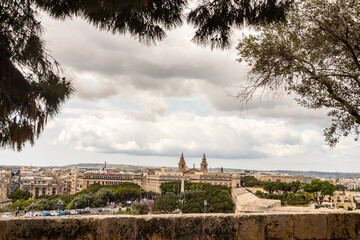Obraz premium Scenic view of Valletta, Malta. A panoramic cityscape of Valletta framed by trees, featuring historic architecture, lush gardens, and church towers under a cloudy sky.