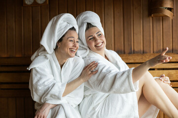 Emphasizing joy, two women laugh together in a sauna while sharing a light-hearted moment as they...