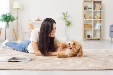  Portrait of young happy smiling female owner playing with her little cute dog lying on floor in the living room at home enjoying leisure together. Cheerful woman with her pet indoors. Animal concept. © Studio Romantic