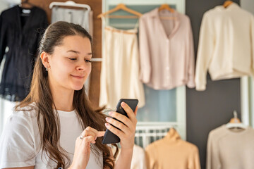 Young woman in a bedroom looking at her smartphone with clothing hanging on a wardrobe behind her. Mobile apps online resale and second-hand fashion, promoting sustainability and smart shopping