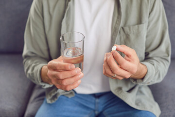 Medicine and health care. Man is sitting on sofa with glass of water and is about to take medicine...