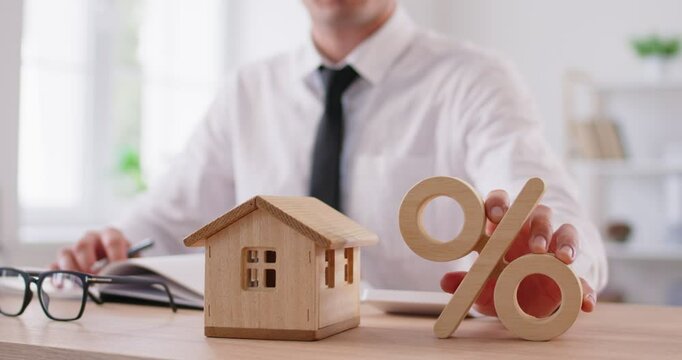 Man in formal clothes sitting at desk in office with wooden toy house and present sign at table. Loan for business investment, mortgage, save money for real estate or buy home concept. 4k video.