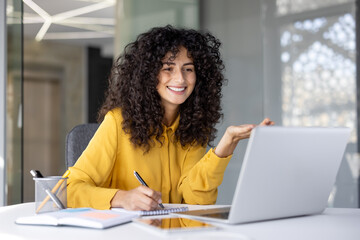 Smiling woman in yellow shirt is attending an online course, taking notes while gesturing in front of her laptop