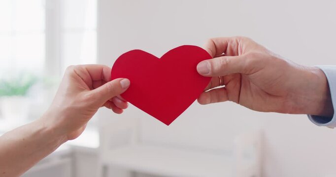 Close up of young married faceless couple holding red paper heart in hands standing living room at home. Love, care and romantic relationships concept. 4k video. Slow motion video.