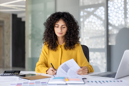 Serious thinking female accountant doing paperwork inside office. Businesswoman recording data, preparing reports, filling out tax forms. Woman with documents and graphs with laptop.