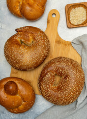 Freshly baked sesame bread and bagels on a wooden cutting board, accompanied by a bowl of sesame seeds and a linen cloth. A rustic bakery composition with golden crusts and soft textures.