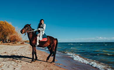 Young smiling asian woman ride on horse on lake shore on summer day