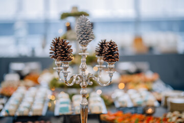 Close-up of a decorative silver candelabrum with pine cones used as centerpieces, with a blurred festive buffet table in the background..