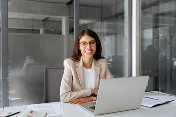 Portrait young it specialist latin hispanic business lady working on laptop pc sitting at desk in modern office smiling at camera. Middle eastern indian woman using computer technology for work online