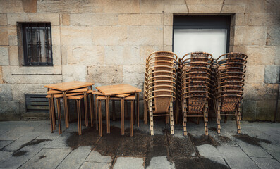 Empty street cafe with stacked wicker chairs and wooden tables against an old stone wall with barred window; wet pavement suggests recent rainfall, rustic urban scene in Europe