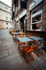 Outdoor cafe with empty wooden tables and chairs on stone-paved sidewalk next to historical stone buildings, big white windows, urban European street, cozy restaurant exterior; Santiago de Compostela