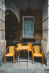 Vintage wooden table with four yellow wicker chairs on stone pavement, set under an old European building archway, framed by stone columns and large window reflecting historic facade