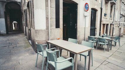 Empty outdoor cafe terrace with modern furniture, turquoise chairs, beige tables, stone pavement, historical European architecture, quiet street atmosphere, no people, daylight; Santiago de Compostela